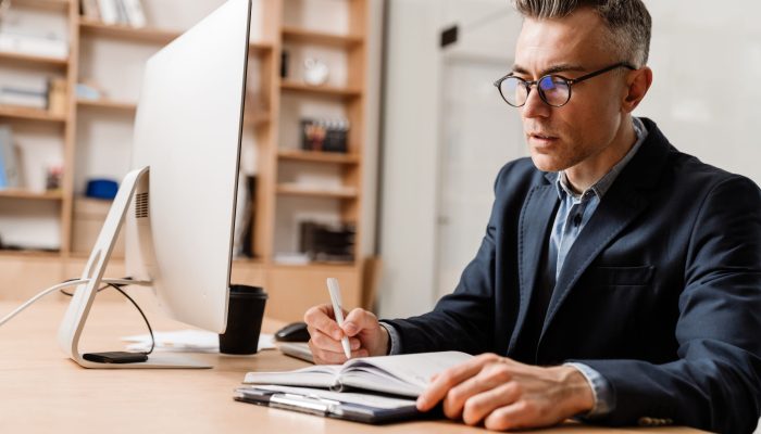 Serious grey man working with computer and planner while sitting at table in office