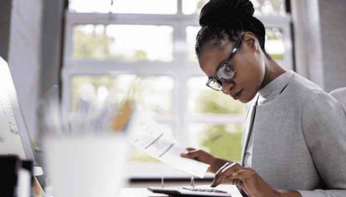 Mulher sentada, em frente à uma mesa, utilizando uma calculadora e um computador, enquanto segura documentos, fazendo cálculos.